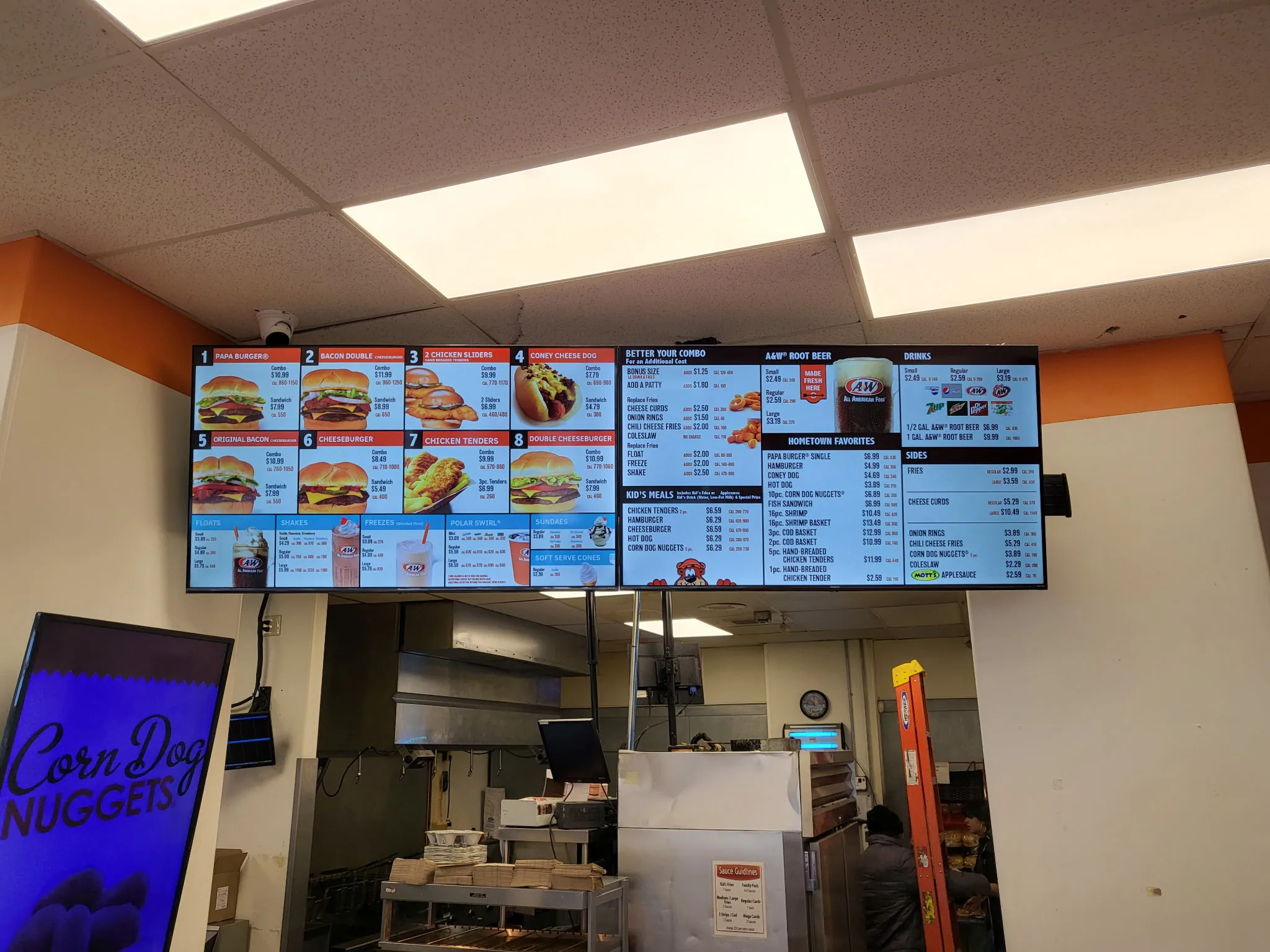 Illuminated digital menu boards mounted above a fast-food service counter, kitchen visible behind