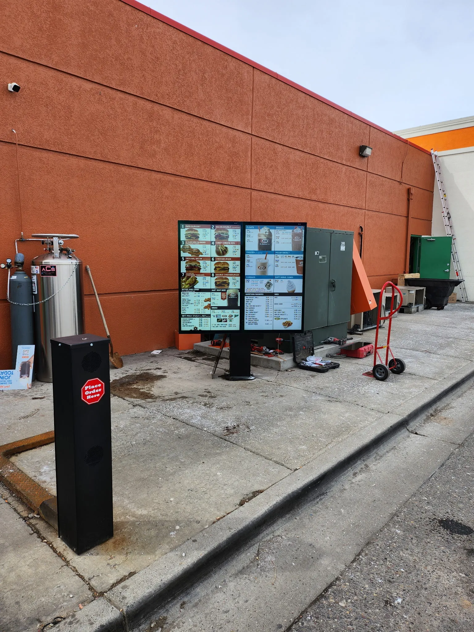 Outdoor drive-thru digital menu screens on a pole beside a red brick restaurant wall, mid-installation